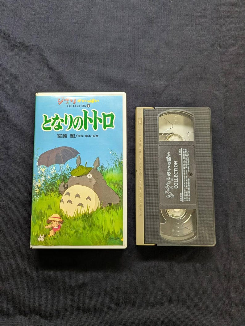 Op de afbeelding: Een VHS-band en hoes met de animatiefilm "My Neighbor Totoro". De hoes is lichtblauw met Japanse tekst en illustraties van Totoro en een jong meisje. De VHS-band is zwart en heeft Japanse tekst.