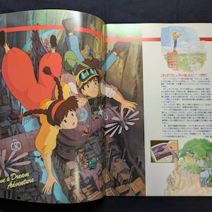 May include: An open book page featuring a colorful anime illustration of two characters falling through the air above a mechanical city. The text Love & Dream Adventure is printed in the corner, alongside Japanese text and smaller inset images.