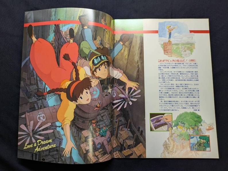 May include: An open book page featuring a colorful anime illustration of two characters falling through the air above a mechanical city. The text Love & Dream Adventure is printed in the corner, alongside Japanese text and smaller inset images.