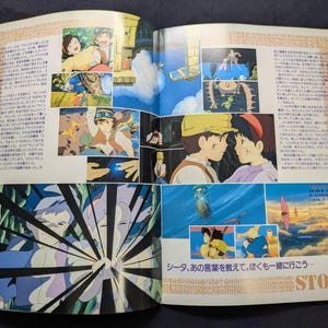 May include: An open book featuring multiple colorful anime-style illustrations from the film Castle in the Sky. The pages contain Japanese text, various scenes of characters in flight and dialogue, and a large graphic panel with black radiating lines.