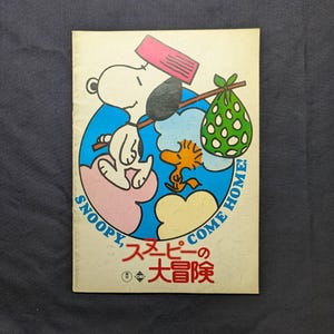 以下が含まれることがあります： 長方形のブックカバー。肩に緑色の水玉模様の風呂敷を棒で担いだスヌーピーと、ウッドストックの漫画イラストが描かれている。テキストには「SNOOPY, COME HOME!」および赤と青で「スヌーピーの大冒険」と記されている。