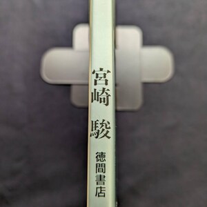 May include: A book spine with Japanese text in black, including the name "Miyazaki" and the publisher "Tokuma Shoten." The book has a light green cover and is held in a gray book holder against a dark background.