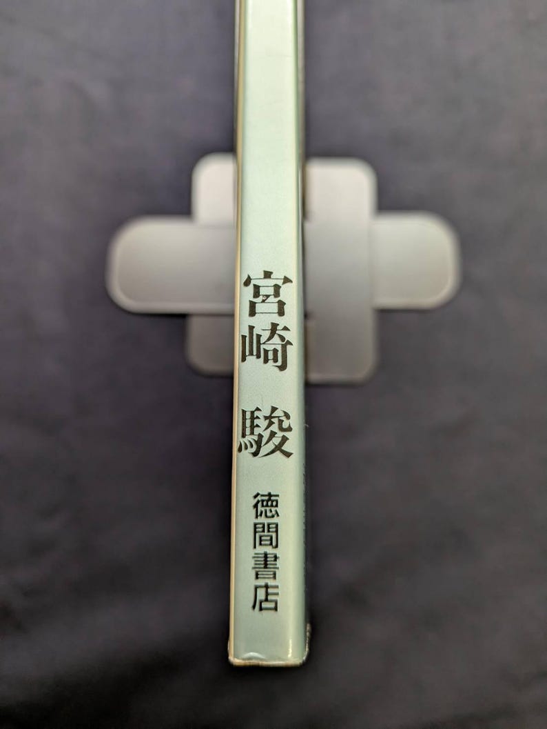 May include: A book spine with Japanese text in black, including the name "Miyazaki" and the publisher "Tokuma Shoten." The book has a light green cover and is held in a gray book holder against a dark background.