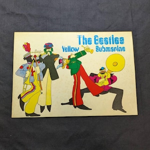 以下が含まれることがあります： 青い文字で「The Beatles Yellow Submarine」と書かれた長方形のプリント。クリーム色の背景に、トロンボーン、トランペット、チューバなどの楽器を演奏する、カラフルなマーチングバンドの制服を着た4人の人物が描かれています。