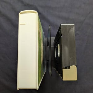 Op de afbeelding: Een witte VHS-videodoos en een zwarte VHS-band worden naast elkaar weergegeven. De videodoos is rechthoekig en heeft een gebogen bovenkant. De VHS-band is zwart met een beige basis.