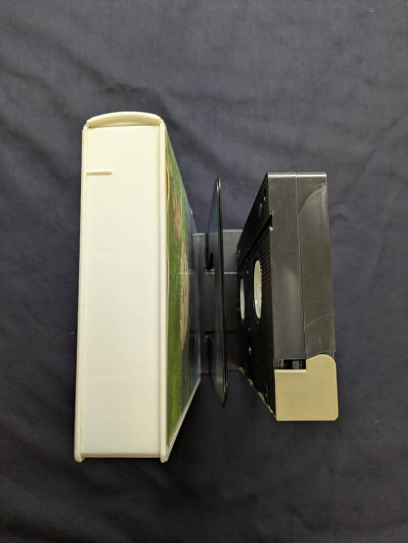 Op de afbeelding: Een witte VHS-videodoos en een zwarte VHS-band worden naast elkaar weergegeven. De videodoos is rechthoekig en heeft een gebogen bovenkant. De VHS-band is zwart met een beige basis.
