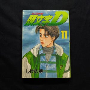 May include: A manga book cover showing a young man in a green and white jacket over a blue plaid shirt. The title "Initial D" is in green and yellow, with the number "11" in yellow. Japanese text is also visible.