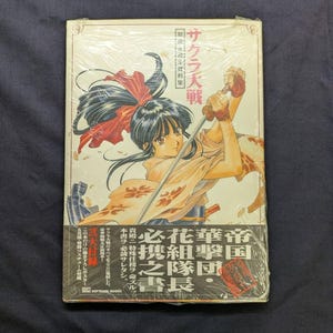 May include: A sealed book with a colorful illustration of a woman with a sword, featuring Japanese text. The cover art showcases a character in a traditional outfit with a red bow. The book is wrapped in clear plastic.
