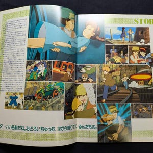 May include: An open book page featuring multiple colorful anime-style illustrations from a film. The layout includes a large central image of a character carrying another, smaller panels showing various scenes, and Japanese text throughout the page.