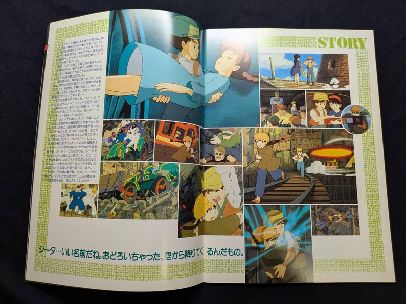May include: An open book page featuring multiple colorful anime-style illustrations from a film. The layout includes a large central image of a character carrying another, smaller panels showing various scenes, and Japanese text throughout the page.