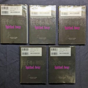 Pode incluir: Cinco c&oacute;pias do livro "Spirited Away" est&atilde;o dispostas sobre uma superf&iacute;cie escura. As capas s&atilde;o escuras com texto japon&ecirc;s e o t&iacute;tulo "Spirited Away" em rosa. Cada livro tem um adesivo branco com texto e um c&oacute;digo de barras.