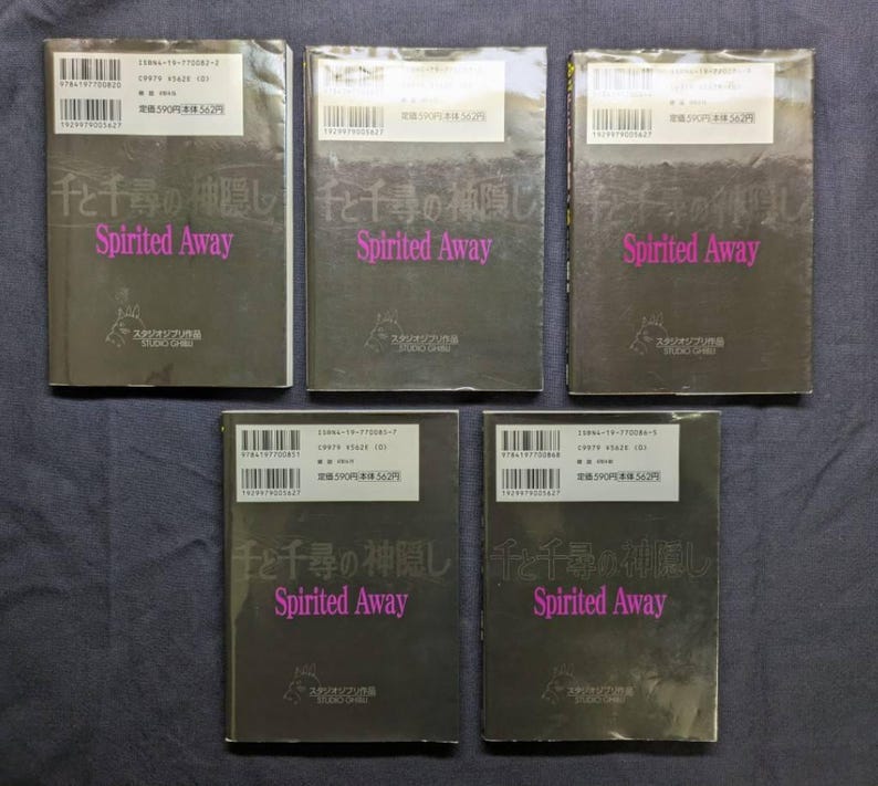 Pode incluir: Cinco c&oacute;pias do livro "Spirited Away" est&atilde;o dispostas sobre uma superf&iacute;cie escura. As capas s&atilde;o escuras com texto japon&ecirc;s e o t&iacute;tulo "Spirited Away" em rosa. Cada livro tem um adesivo branco com texto e um c&oacute;digo de barras.