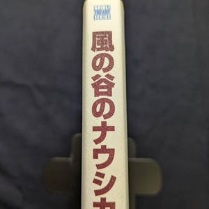 Pu&ograve; includere: Dorso di libro con testo giapponese in rosso e le parole "Ghibli The Art Series" in blu. Il libro &egrave; bianco sporco e tenuto in un leggio nero. Il testo giapponese &egrave; orientato verticalmente.