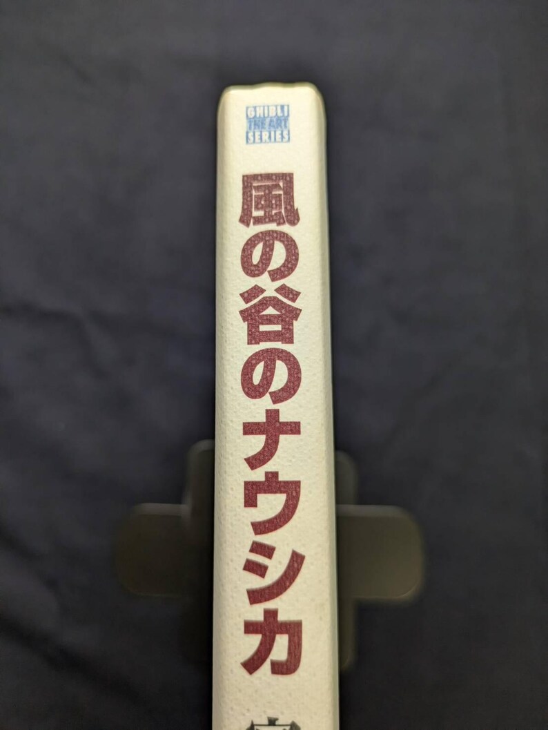 Pu&ograve; includere: Dorso di libro con testo giapponese in rosso e le parole "Ghibli The Art Series" in blu. Il libro &egrave; bianco sporco e tenuto in un leggio nero. Il testo giapponese &egrave; orientato verticalmente.