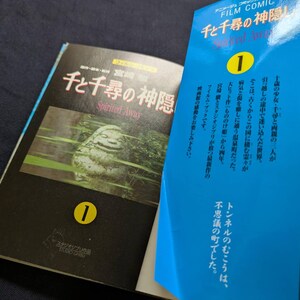 Pode incluir: Um livro aberto e um livro azul parcialmente vis&iacute;vel. O livro aberto tem uma capa preta com texto japon&ecirc;s e o t&iacute;tulo "Spirited Away" em ingl&ecirc;s. O livro azul tem texto japon&ecirc;s e o n&uacute;mero "1" em um c&iacute;rculo amarelo.