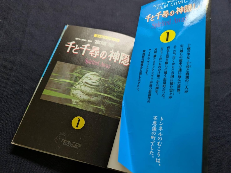 Pode incluir: Um livro aberto e um livro azul parcialmente vis&iacute;vel. O livro aberto tem uma capa preta com texto japon&ecirc;s e o t&iacute;tulo "Spirited Away" em ingl&ecirc;s. O livro azul tem texto japon&ecirc;s e o n&uacute;mero "1" em um c&iacute;rculo amarelo.