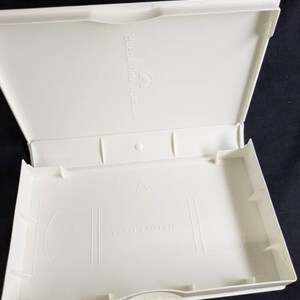 Op de afbeelding: Een open, cr&egrave;mekleurige VHS-videocassettecase. De case is rechthoekig met een scharnierend deksel. De binnenkant heeft een gegoten ontwerp om een VHS-band vast te houden, met de woorden "VIDEO CASSETTE" onderaan gedrukt. De case is gemaakt van plastic.
