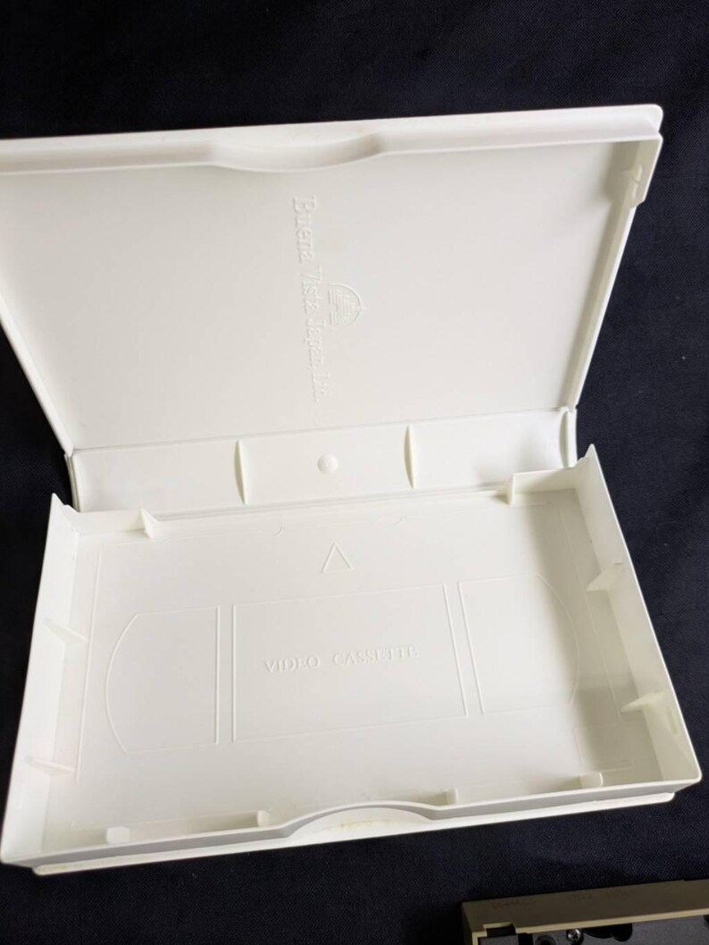 Op de afbeelding: Een open, cr&egrave;mekleurige VHS-videocassettecase. De case is rechthoekig met een scharnierend deksel. De binnenkant heeft een gegoten ontwerp om een VHS-band vast te houden, met de woorden "VIDEO CASSETTE" onderaan gedrukt. De case is gemaakt van plastic.