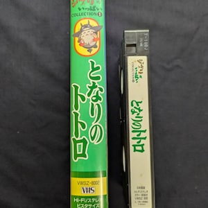 Op de afbeelding: Twee VHS-banden, een lange en een korte, met Japanse tekst en illustraties. De lange band is groen met de titel "Mijn buurman Totoro" en een stripfiguur. De kortere band is zwart met witte tekst en dezelfde titel.