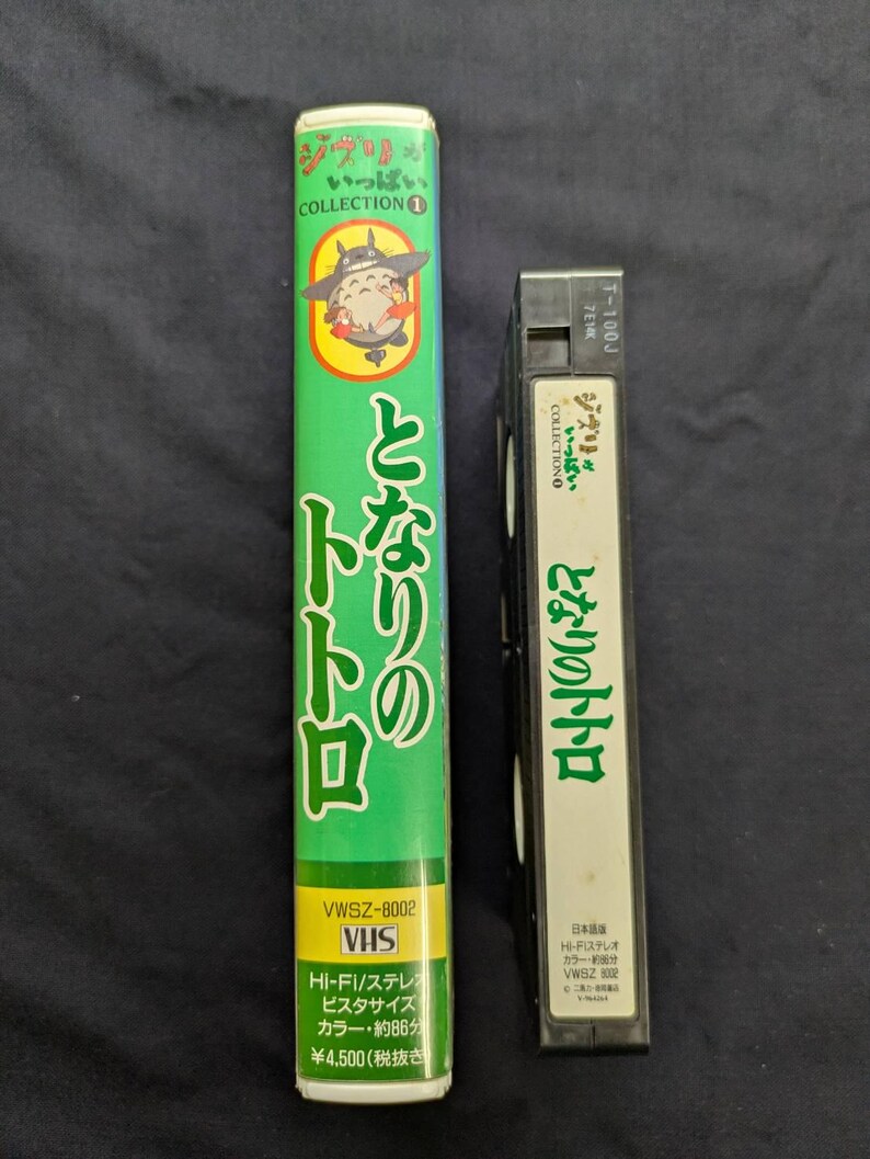 Op de afbeelding: Twee VHS-banden, een lange en een korte, met Japanse tekst en illustraties. De lange band is groen met de titel "Mijn buurman Totoro" en een stripfiguur. De kortere band is zwart met witte tekst en dezelfde titel.
