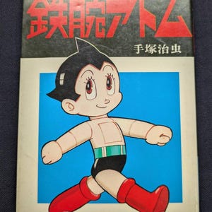 May include: A vintage comic book featuring Astro Boy. The cover has the title "Mighty Atom" in English and Japanese characters, with a sun graphic. Astro Boy is depicted in a running pose, wearing red boots, black shorts, and a green belt. The number "2" is visible.