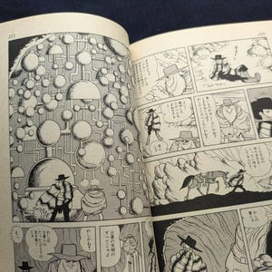 May include: Open book with black and white manga illustrations. The panels depict a science fiction theme with characters in hats and coats, and a complex network of spheres and lines. Text is present in Japanese.