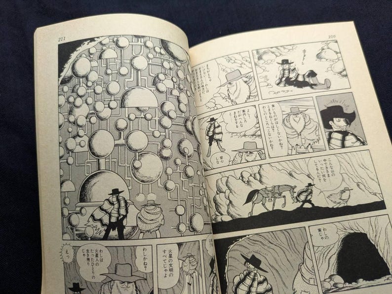 May include: Open book with black and white manga illustrations. The panels depict a science fiction theme with characters in hats and coats, and a complex network of spheres and lines. Text is present in Japanese.