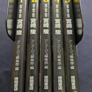 Pode incluir: Um conjunto de cinco livros japoneses com lombadas pretas. Cada livro tem um n&uacute;mero diferente em amarelo ou vermelho e texto japon&ecirc;s. Os livros est&atilde;o em um suporte preto.