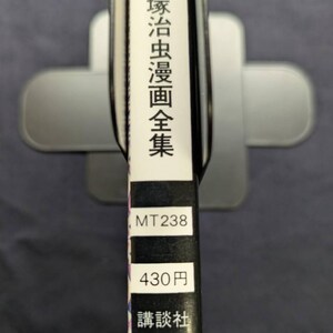 May include: A book spine with Japanese text and the title "Tezukamushi Manga Zenshu". The spine also includes the numbers "MT238" and "430円", along with the publisher's name. The book is held in a silver book stand.
