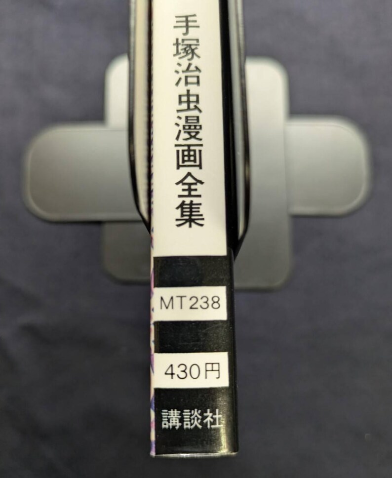 May include: A book spine with Japanese text and the title "Tezukamushi Manga Zenshu". The spine also includes the numbers "MT238" and "430円", along with the publisher's name. The book is held in a silver book stand.