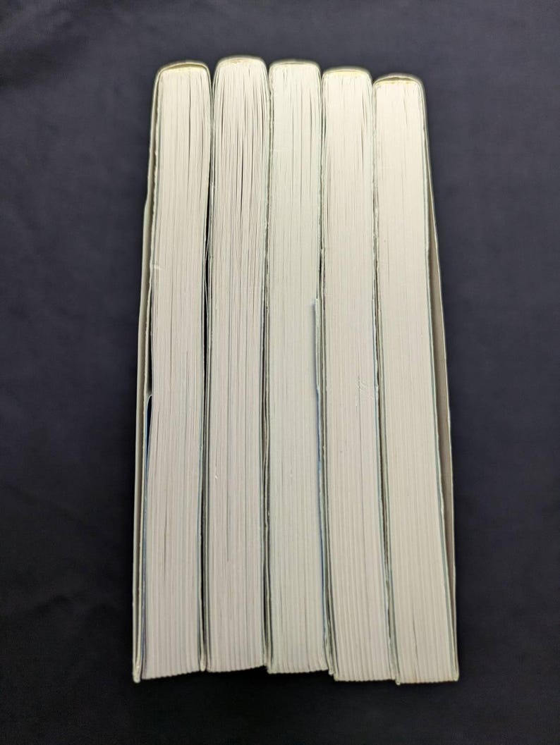 Pode incluir: Uma pilha de cinco livros com p&aacute;ginas brancas e bordas verde claro. As lombadas n&atilde;o s&atilde;o vis&iacute;veis e os livros est&atilde;o dispostos verticalmente sobre um fundo escuro. As p&aacute;ginas est&atilde;o ligeiramente amareladas.