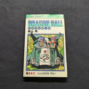 May include: A Dragon Ball manga volume featuring a green race car with two characters. The cover has the title "DRAGON BALL" in large, colorful letters. The text "JUMP COMICS" is at the top. The volume is labeled "Volume 32".