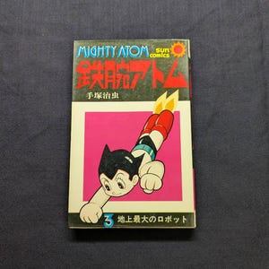 May include: A vintage comic book titled "Mighty Atom" featuring a cartoon character in a flying pose. The cover has a pink background with Japanese text and the number 3. The comic book is rectangular and has a colorful design.