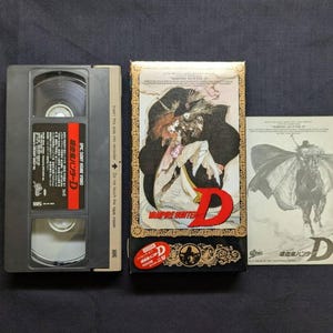 以下が含まれることがあります： 暗い表面に表示された「Vampire Hunter D」のヴィンテージVHSテープ、ボックス、インサート。ボックスには、大きな赤い「D」が付いた詳細なイラストが描かれています。テープには日本語のテキストと赤いラベルが付いています。