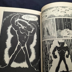 May include: A black and white comic book illustration featuring a muscular figure with a horned head and a star on the chest. The figure is flexing its arms. The comic book is open, displaying multiple panels.