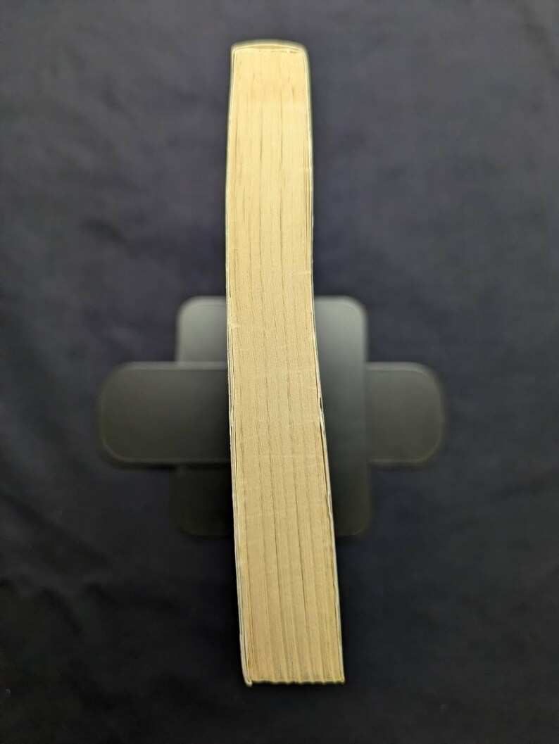 May include: A hardcover book with beige-colored pages, held upright by a black book stand. The book's spine is visible, showing the stacked pages. The background is a dark, solid color.