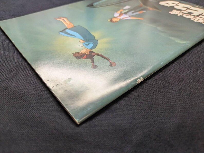 May include: A rectangular book cover featuring a teal background with an illustration of a person in a blue dress falling through the air, accompanied by another figure above, with Japanese text visible in the upper right corner of the cover.