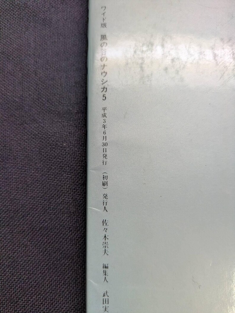 May include: A book with Japanese text. The cover is light blue with Japanese text in black. The text includes "ワイド版 風の谷のナウシカ5" and the date "平成3年6月30日". The book is on a dark gray surface.