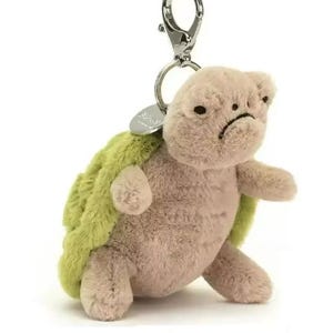 May include: A soft toy turtle keychain in tan and green. The turtle has a sad expression and a silver metal clip. The plush toy is designed with a green shell and a tan body.