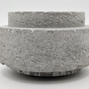 May include: A gray concrete object with a rough, textured surface. The object has a cylindrical shape with a smaller, raised circular top. The base is also circular, giving it a tiered appearance. The overall design is minimalist and industrial.