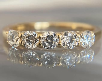 May include: A gold ring with five round diamonds set in a row. The diamonds are clear and sparkling, set in a yellow gold band. The ring's design is simple, with the diamonds being the focal point. The ring is reflecting on a shiny surface.