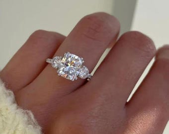 May include: A dazzling engagement ring featuring a large, cushion-cut central diamond and smaller side stones. The ring is set in a silver band and is shown on a hand.