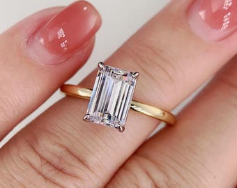 May include: A gold ring featuring a large, rectangular, clear gemstone. The ring is set on a gold band and is held by four prongs. The gemstone is faceted and reflects light.