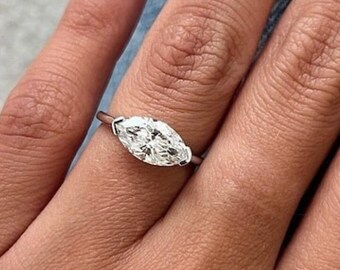 May include: A silver ring featuring a marquise-cut diamond. The diamond is set horizontally. The ring is on a finger, with a blurred denim background. The ring's design is simple and elegant.
