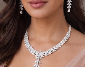 May include: A dazzling diamond necklace and earrings set. The necklace has a floral design with a teardrop pendant. The earrings are a delicate leaf-shaped design. The jewellery is set against a soft, peach-coloured background.