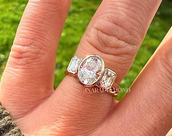 May include: A gold ring featuring a central oval-cut stone in a bezel setting, flanked by two smaller rectangular-cut stones on either side. The band is polished gold, and the text INARADIAMOND is visible on the metal setting near the stones.