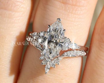 May include: A silver-coloured ring set featuring a large marquise-cut centre stone surrounded by smaller round and marquise-cut accent stones. The band is curved and adorned with pavé-set stones, with the text INARADIAMOND visible on the metal.