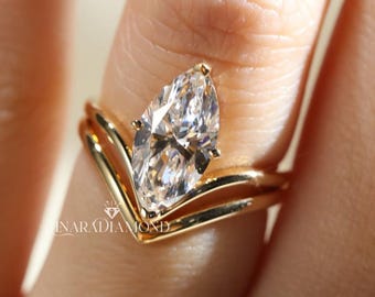 May include: A gold ring set featuring a marquise-cut clear gemstone held by prongs, paired with a matching V-shaped gold band. The metal has a polished finish, and the text INARADIAMOND is visible on the band near the base of the setting.