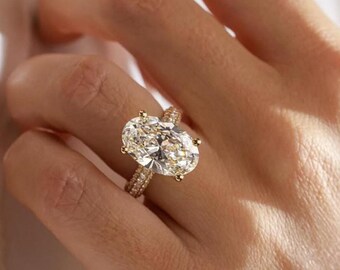 May include: Close-up of a gold ring featuring a large oval-cut diamond. The ring has a pave band and is on a hand. The diamond is clear and sparkling, reflecting light.