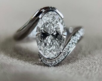 May include: A silver-toned ring featuring a large, oval-cut centre stone held by a bypass-style setting. One side of the band is smooth and polished, while the other side is encrusted with small, round, clear stones. The text ANARADIAMONDS is visible.